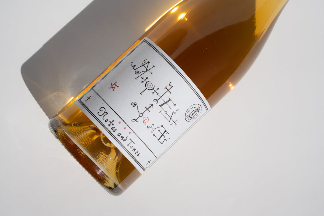 A close-up of the 2022 Notes & Tones - The Wine bottle by Broc Cellars lies on a white surface. Its label features abstract symbols and decorative fonts. The golden amber-tinted bottle casts a shadow to the right, evoking an exclusive vibe.