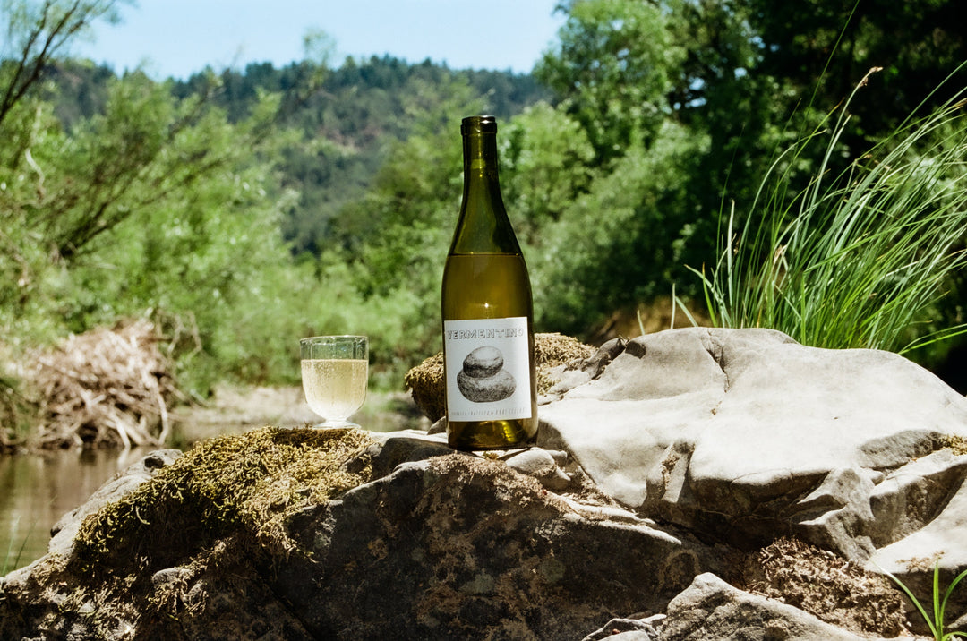 Six bottles of wine with unique labels and three glasses filled with red liquid are arranged on dry grass outdoors, with sunlight and forested hills in the background.