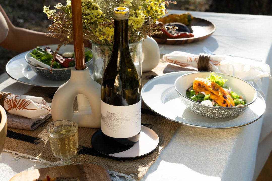 A dark wine bottle with a white label featuring an illustration of twisted roots or branches, set against a light background with plant shadows cast around it.