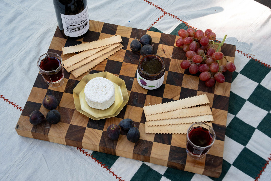 A wooden board with a round of soft cheese cut into wedges on a gold plate, a jar of fig spread, crackers, fresh figs, and grapes.