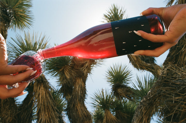A bottle of Broc Cellars 2022 Love Red, made from organically farmed grapes, is poured into a glass. With a backdrop of palm trees and blue sky, one hand holds the bottle while the other holds the glass.