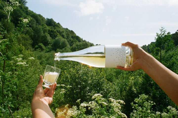 Outdoors beneath a clear sky, someone pours Broc Cellars 2024 Love Sparkling Chenin, a light-colored wine, from the bottle into a glass with green trees and bushes in the background.