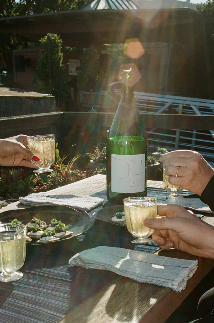 Three people hold small glasses of Broc Cellars 2024 Sparkling Chenin Magnum around an outdoor table with a bottle and plates of food, sunlight, and greenery—perfect for enjoying the flavors of Paso Robles’ Shell Creek Vineyard.