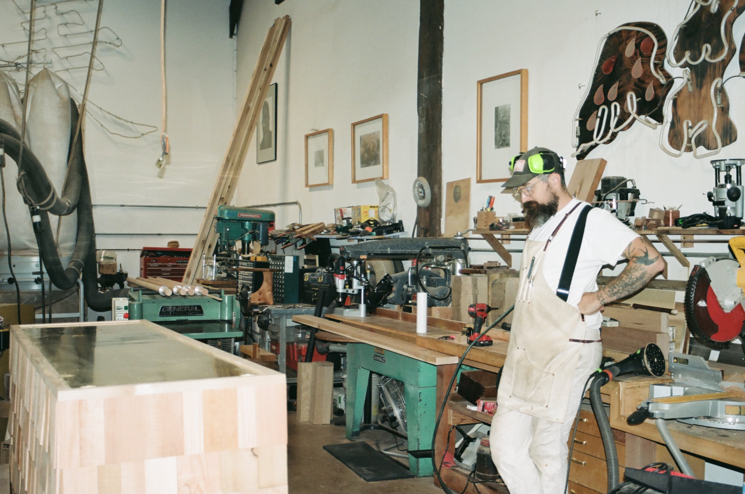 A person wearing a white shirt, apron, and ear protection stands in a woodworking shop, surrounded by benches, tools, machinery, and framed art on the walls.