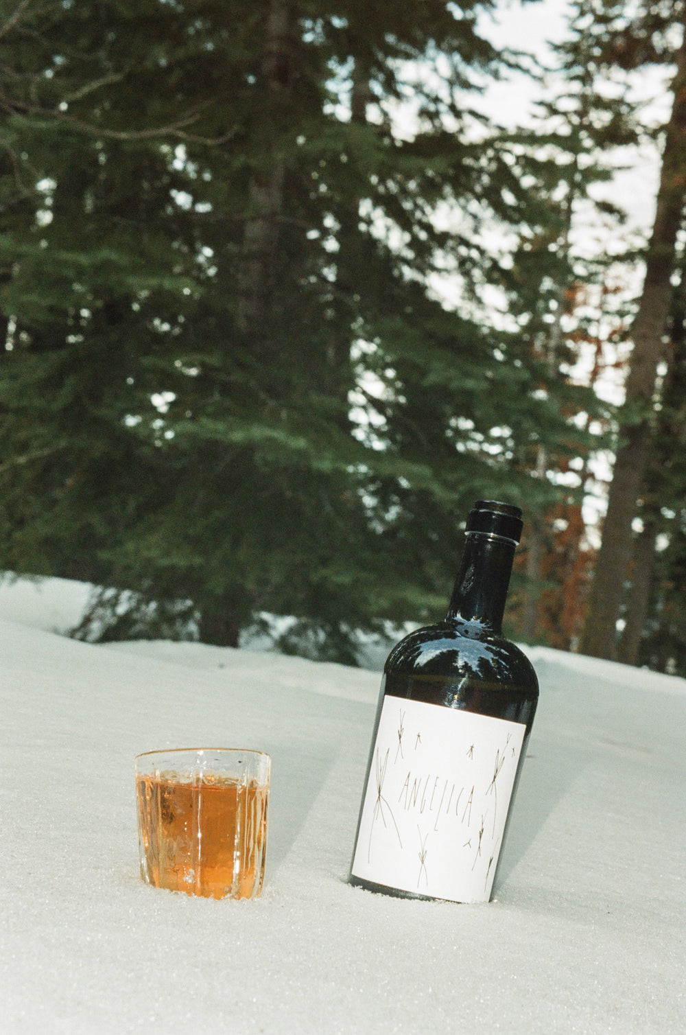 A glass of amber-colored 2020 Angelica with a dark bottle labeled Broc Cellars sits on snow, flanked by wine glasses and evergreen trees in the background.