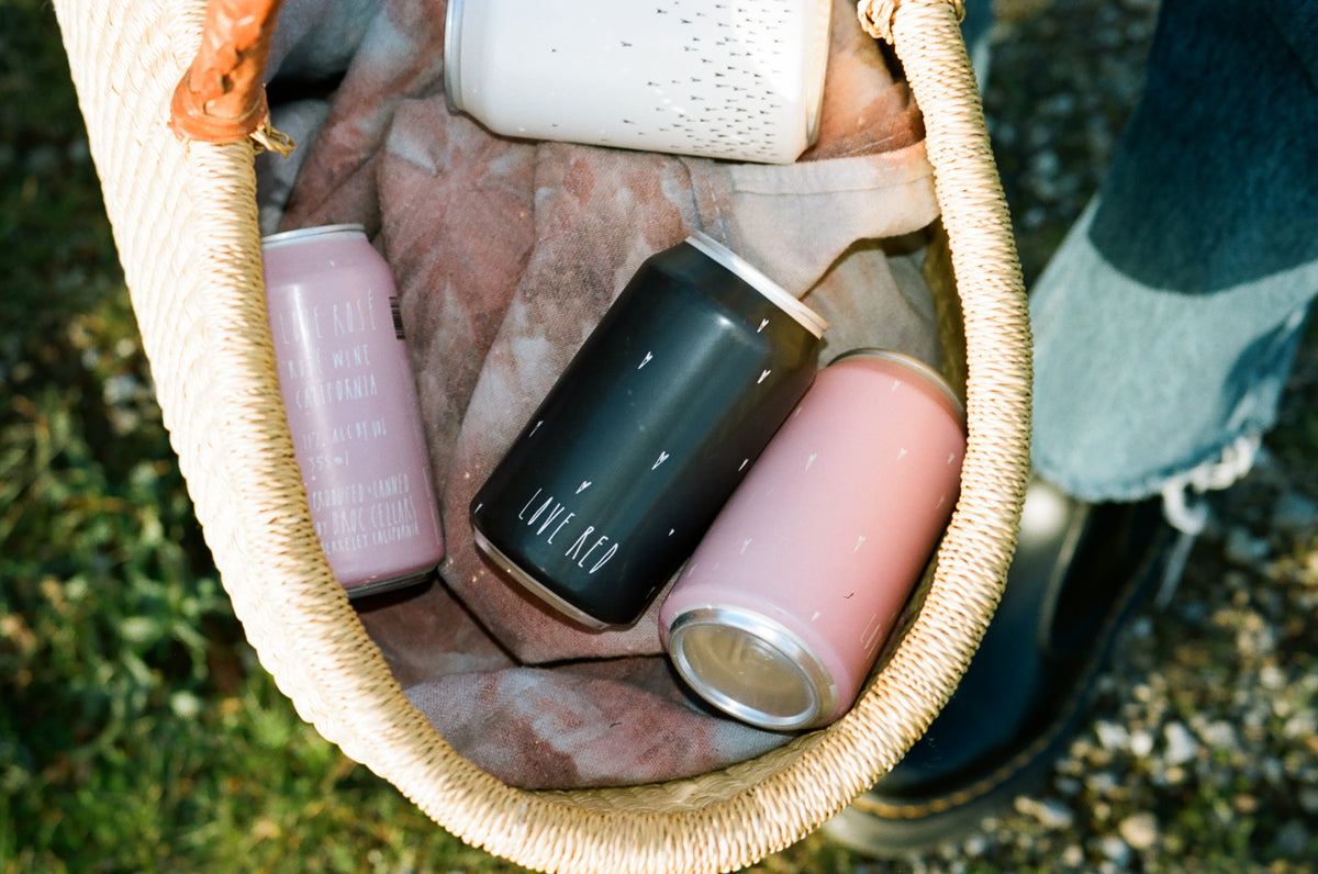 A woven basket with a cloth lining holds four cans of drinks in pink, black, and white, alongside a persons foot wearing a black shoe and denim pant. The basket is on grass.
