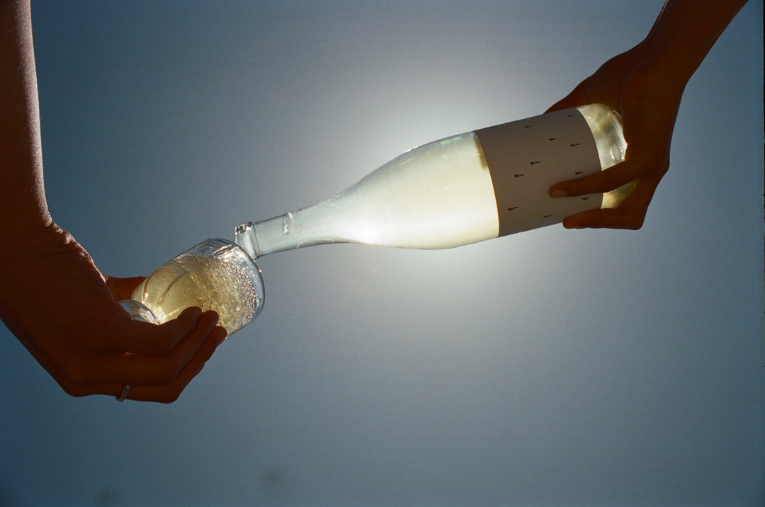 A hand pours Broc Cellars’ 2024 Love White, a sparkling Rhône blend, from a clear bottle into a glass beneath sunlight and a blue sky.