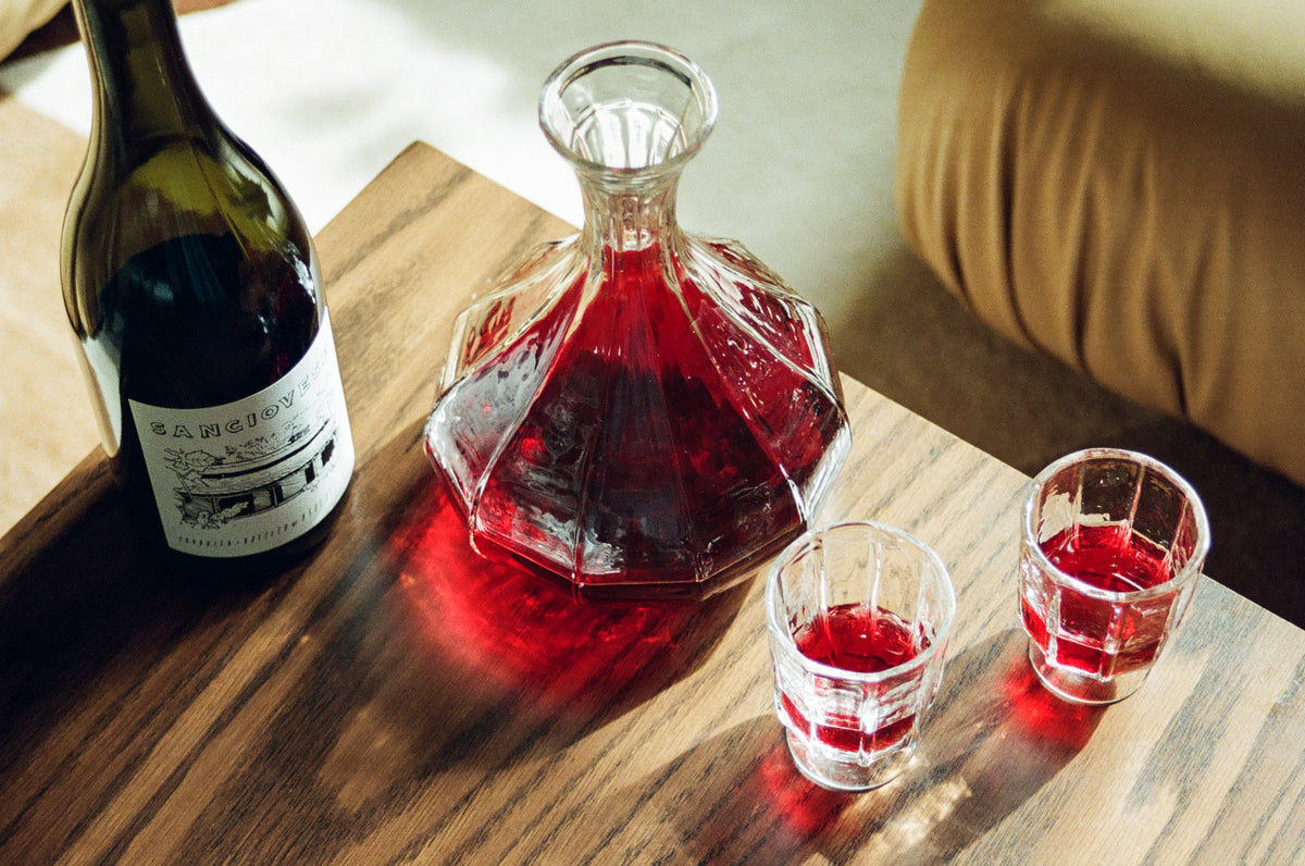 A bottle of red wine, a crystal decanter filled with red wine, and two small glasses with red wine are placed on a wooden table. Sunlight casts reflections and shadows on the surface.