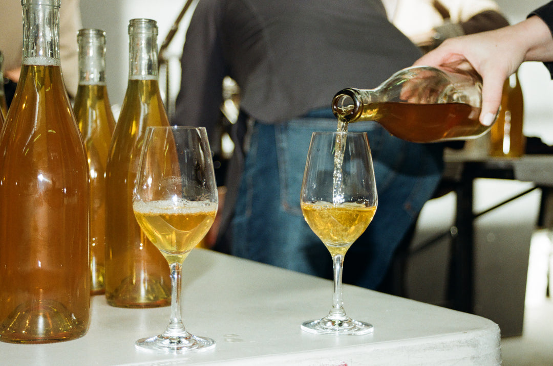 Someone pours golden-colored 2022 Notes &amp; Tones - The Wine by Broc Cellars into a glass on a table, surrounded by more bottles and another poured glass, with people visible in the background.