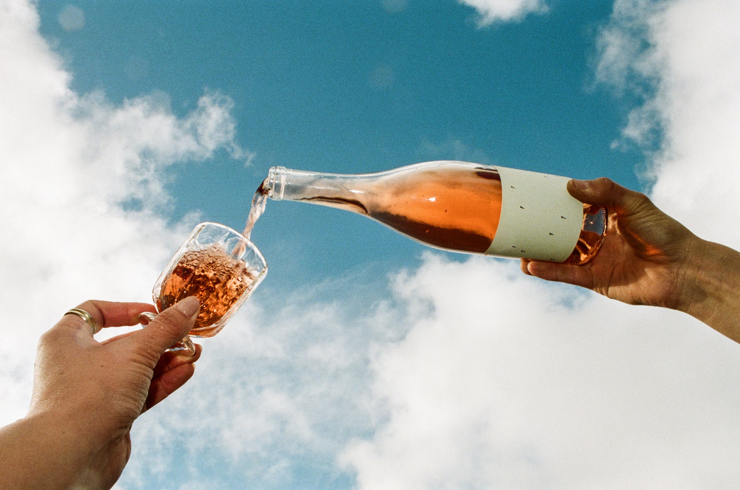 A person pours Broc Cellars 2025 Love Rosé from a clear bottle into a glass held by another, with blue sky and clouds in the background.