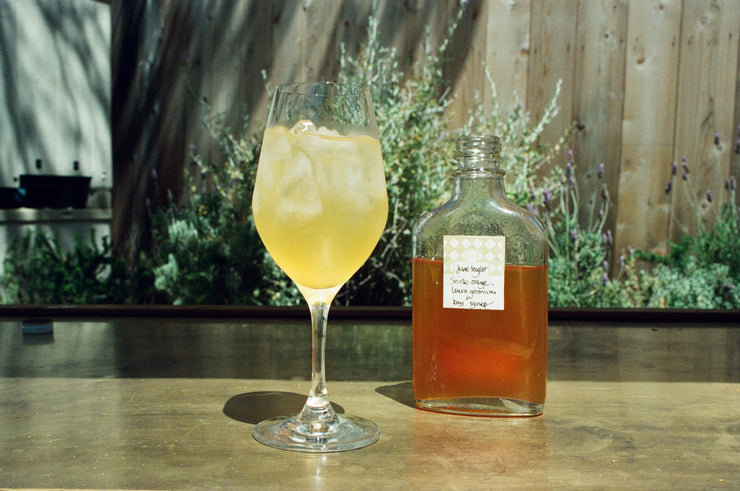 A glass of iced yellow liquid, evoking Broc Cellars Spritz Pack with Clementine + Lemon Thyme Syrup, rests atop wood near a labeled Pét-Nat bottle. A rustic wooden fence and vibrant green foliage enhance the setting.