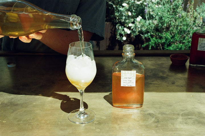 Someone pours Broc Cellars Spritz Pack w/ June Taylor Seasonal Fruit Syrup into a wine glass with ice. A clear bottle with amber syrup and a handwritten label sits beside the glass, with greenery in the background.