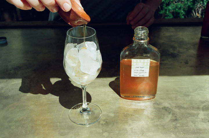 A hand pours liquid into a wine glass filled with ice, beside a square bottle of Broc Cellars&