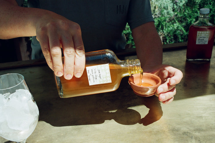 A person pours a brown liquid from a labeled Broc Cellars bottle into a clay cup, reminiscent of enjoying the Spritz Pack with Clementine + Lemon Thyme Syrup outdoors. A glass with ice is nearby, and another bottle is in the background amidst greenery.