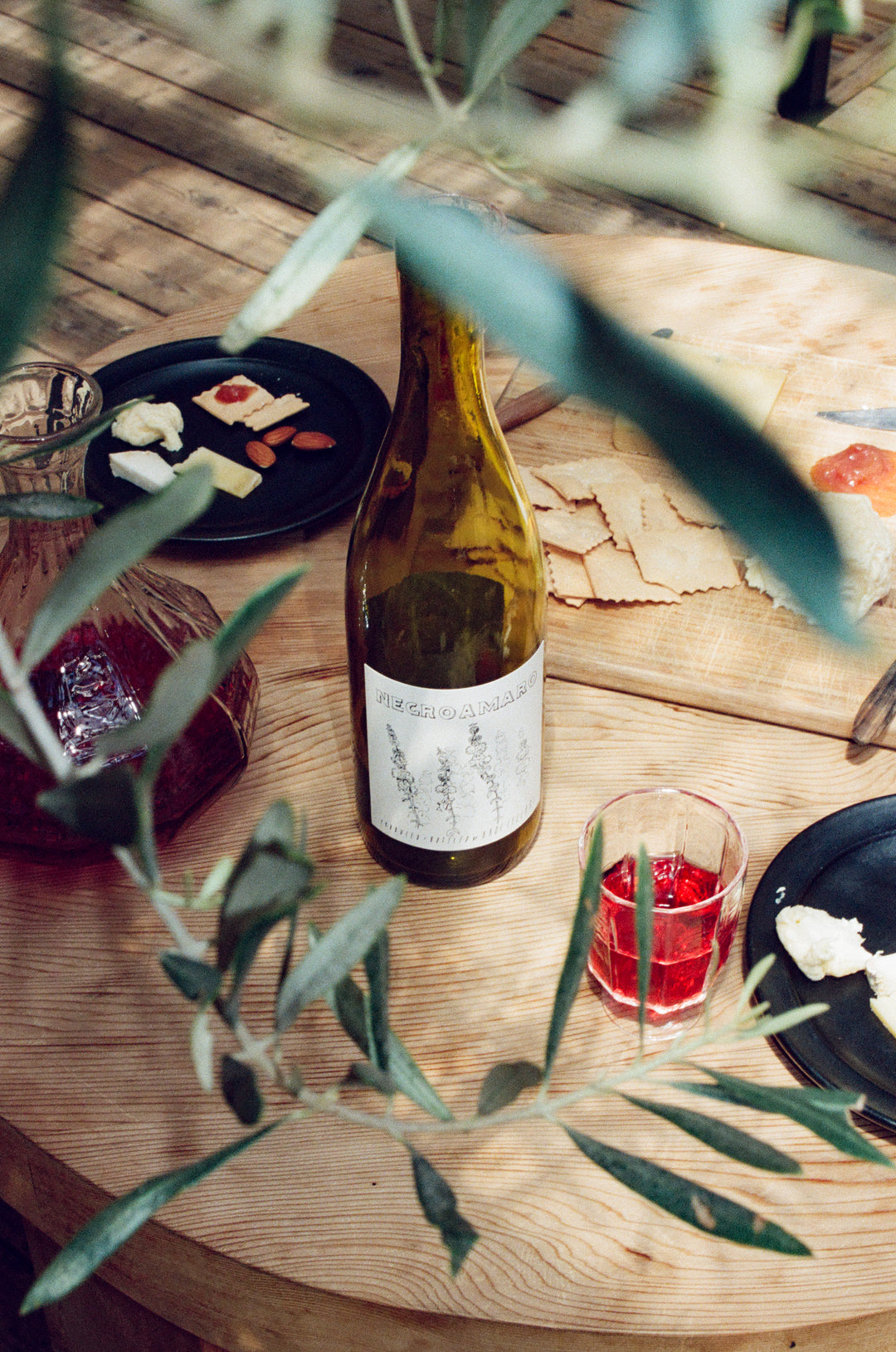 A casual outdoor table features Broc Cellars 2024 Negroamaro, a glass of red wine, a decanter, plates of cheese, crackers, and fruit. A leafy branch partially obscures the view.