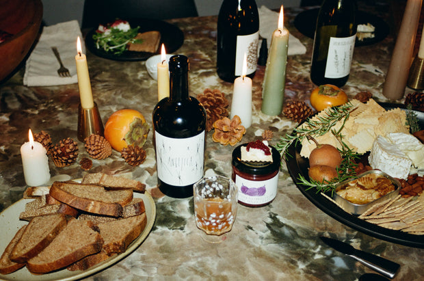 A table is adorned with a glass of Broc Cellars Angelica wine, candles, bread, and assorted cheeses. Crackers, persimmons, and pinecones add charm. June Taylor Mission Fig + Angelica Jam beautifully pairs with the spread. Wine bottles and a salad plate in the background enhance the warm atmosphere.