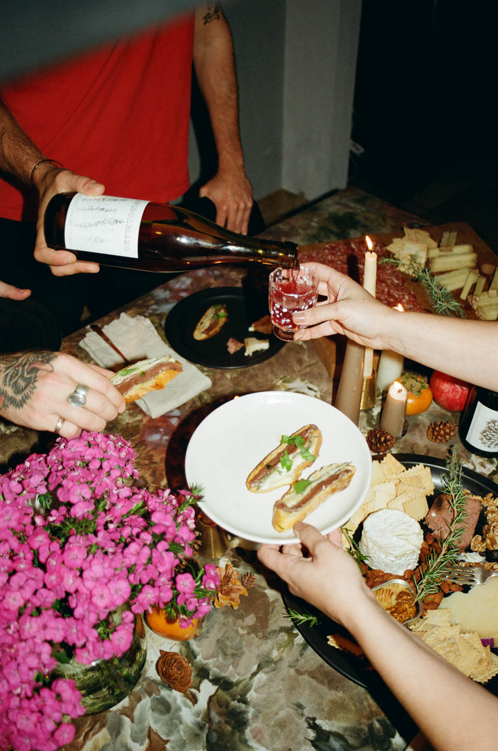 Someone pours Broc Cellars 2024 Negroamaro into a glass held across a table set with flowers, candles, plates of food, cheeses, and snacks. Multiple hands serve and hold food in this vibrant gathering scene.