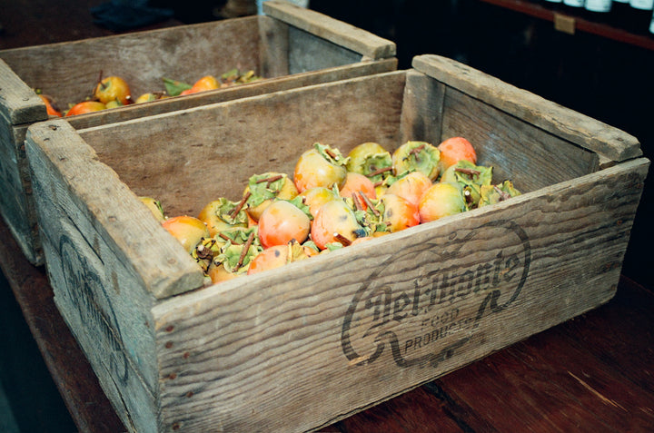A wooden crate filled with ripe persimmons sits on a dark wooden table, evoking ingredients used in Broc Cellars’ June Taylor Persimmon Butter. Another crate is partly visible behind, both boasting vintage-style text on their sides.