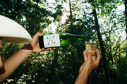 A person in a white hat pours Broc Cellars 2024 Arneis, from a green Certified Organic bottle, into a clear glass outdoors among trees and sunlight.