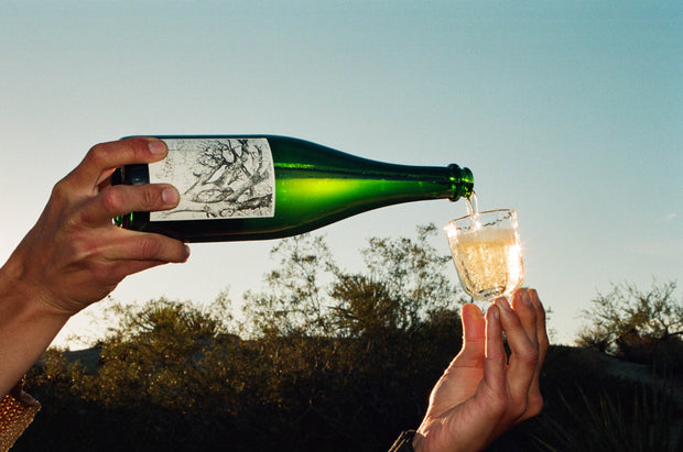 A person pours Broc Cellars 2024 Sparkling Erbaluce from a green bottle into a glass outside, with trees and a clear sky in the background.