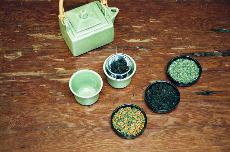 The Broc Cellars Mothers Day Pack is displayed on a wooden surface with a green ceramic teapot, teacup, strainer, and four small bowls filled with assorted loose leaf teas and herbs.