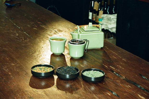 A Broc Cellars Mothers Day Pack is displayed on a wooden table with a green teapot, two teacups (one with tea, one with strainer), and three small bowls of loose leaf tea. Bottles and folded towels appear in the background.