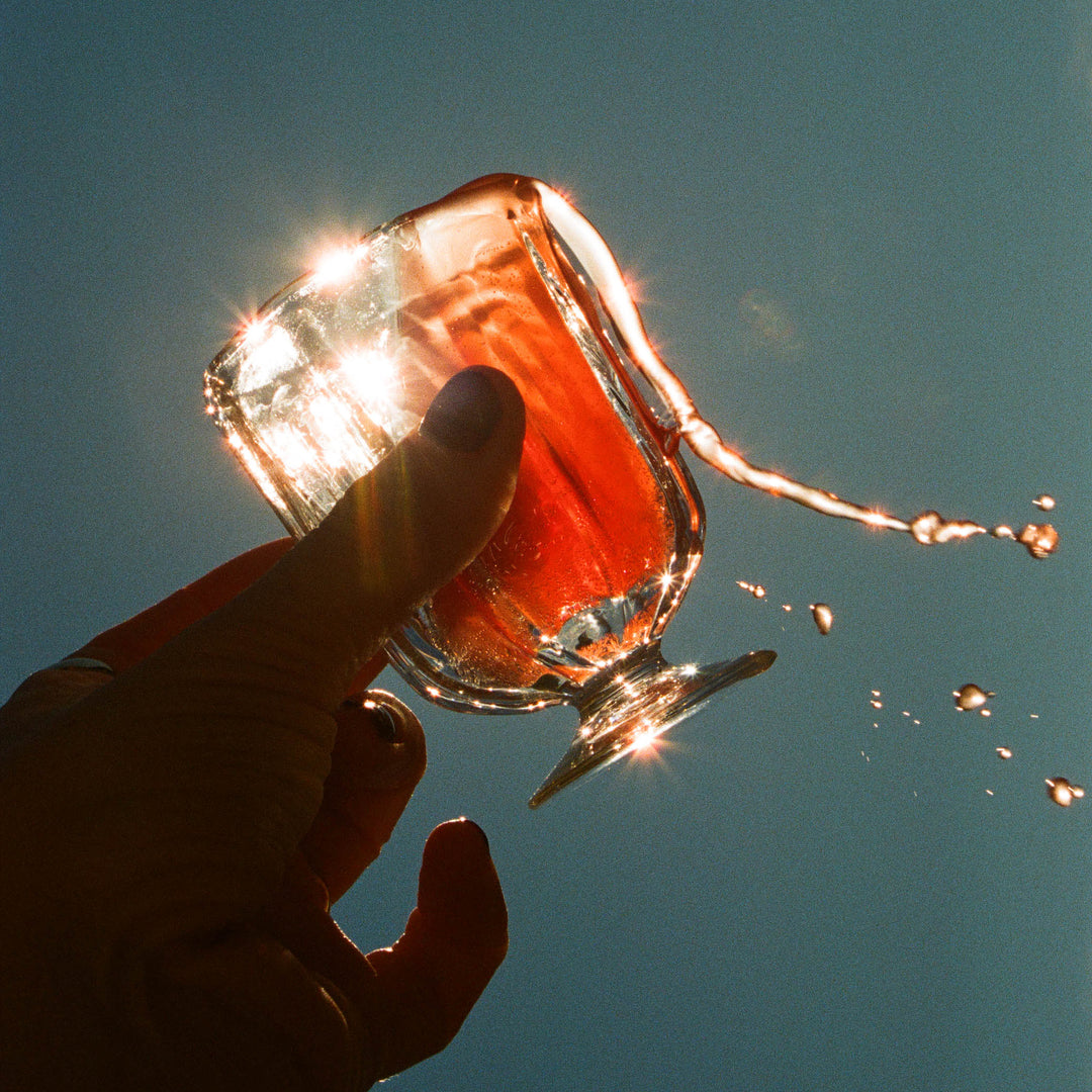A hand holds a glass of red liquid tilted against a clear blue sky, causing the drink to spill out in midair. Sunlight reflects off the glass and liquid, creating bright, star-like flares.