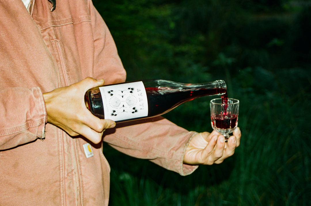 A person in a pinkish jacket pours 2023 Ayay! by Broc Cellars into a small glass. The green, blurred background suggests an outdoor setting, adding to the elegance of this coferment experience.