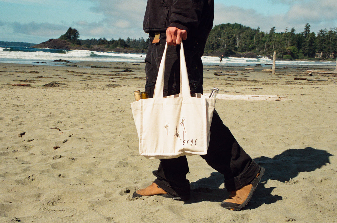 A person in dark clothing and brown boots walks on a sandy beach carrying the Broc Bottle Tote by Broc Cellars, with bottle compartments. The ocean and a forested shoreline are visible in the background.