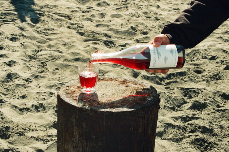 Someone pours Broc Cellars 2023 Amore Blendo into a clear plastic cup on a wooden stump. The scene unfolds on a sandy beach in daylight, echoing the rustic charm of Piemonte.