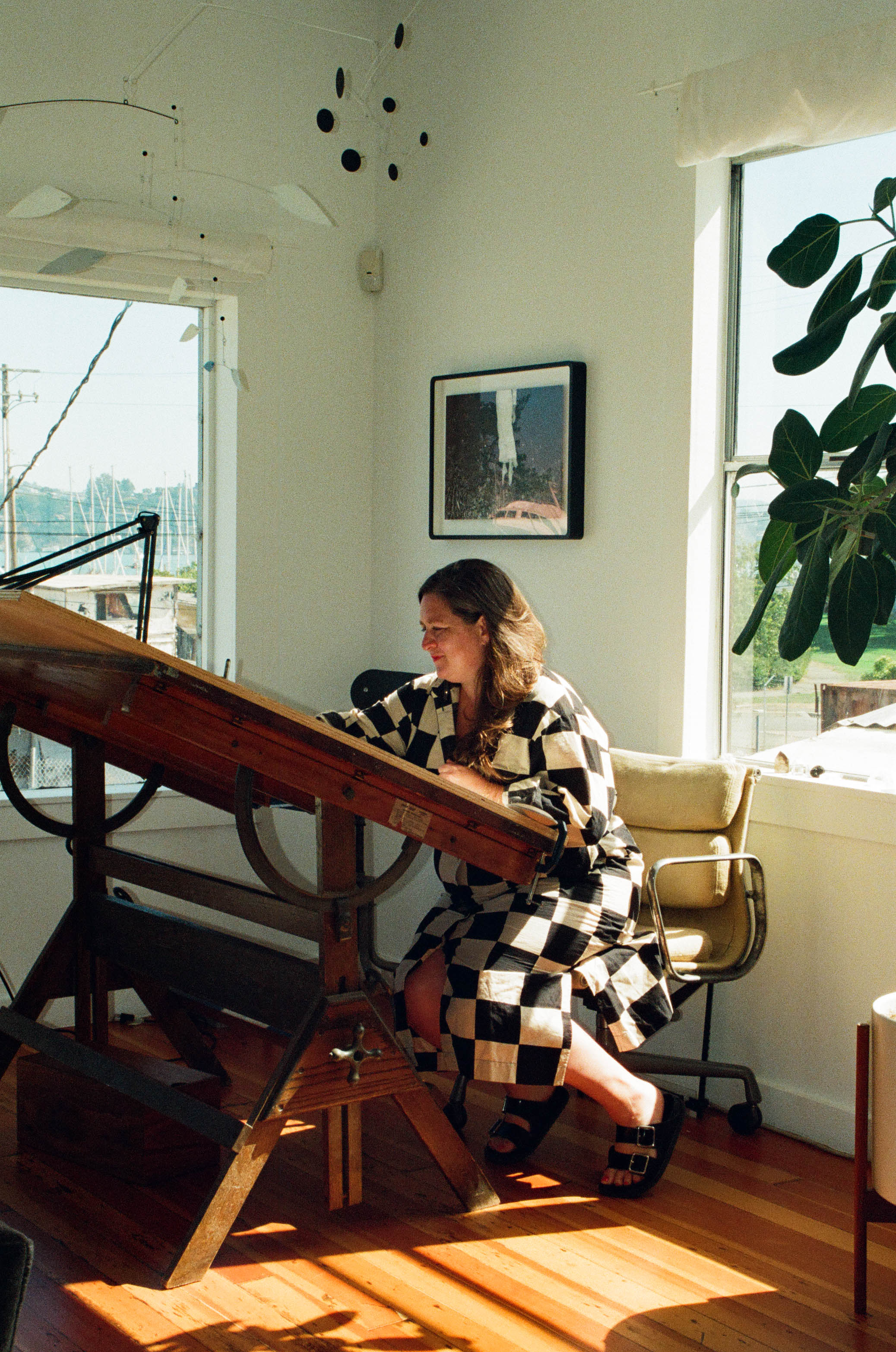A person wearing a black and white checkered outfit sits at a large wooden drafting table in a bright room with large windows, a houseplant, and framed artwork on the wall.