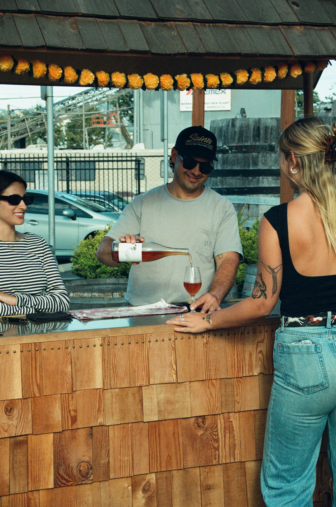 A man wearing a cap pours a glass of rosé wine for a woman at an outdoor wooden bar, while another woman in sunglasses stands nearby smiling. A car and a fence are visible in the background.