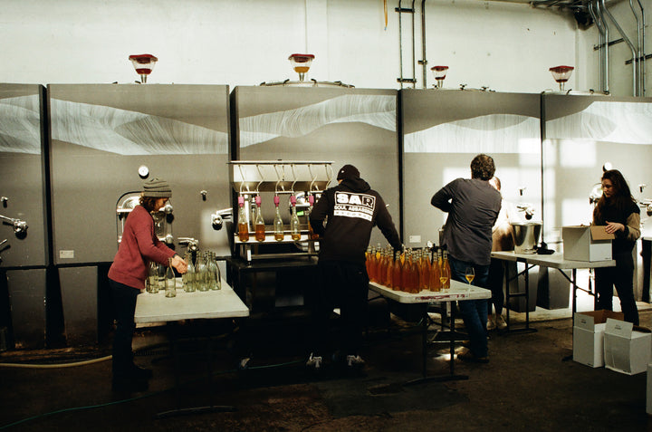 In Broc Cellars beverage facility, four workers diligently fill, cap, and label bottles for the Notes &amp; Tones V.2 - The Album and Wine Set. Amidst tables of equipment and bottles, the industrial atmosphere buzzes with their collaborative efforts to infuse a Broc Cellars touch into every step.