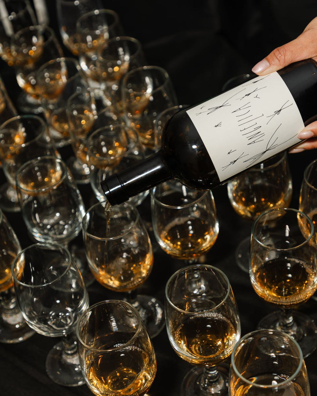 A hand pours white wine from a bottle with a minimalist label into one of several clear glasses, some already filled, arranged on a black surface.
