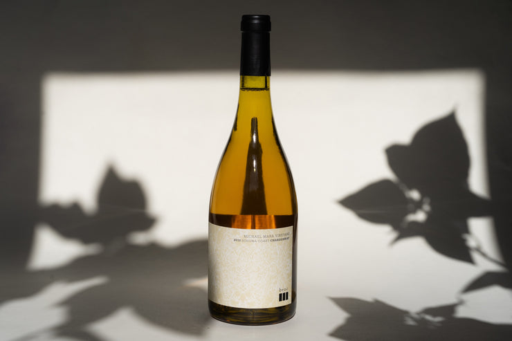 A bottle of 2010 Michael Mara Chardonnay from Broc Cellars rests against a light backdrop, where leaf shadows create a striking contrast. Its light-colored label with simple text embodies the spirit of the Sonoma Coast.