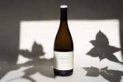 A bottle of 2016 Michael Mara Chardonnay by Broc Cellars sits elegantly on a surface, its label highlighting origins from Sonoma Countys Michael Mara Vineyard. Leaf shadows in the softly lit background enhance the wines refined allure with an alcohol content of 13.4%.