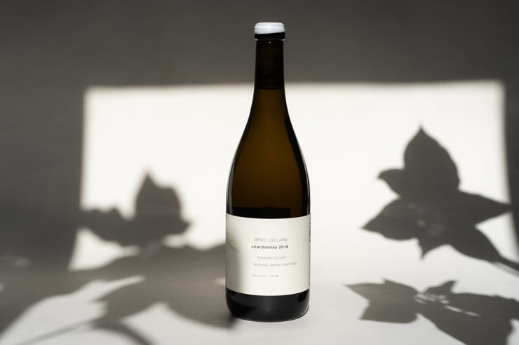 A bottle of 2016 Michael Mara Chardonnay by Broc Cellars sits elegantly on a surface, its label highlighting origins from Sonoma Countys Michael Mara Vineyard. Leaf shadows in the softly lit background enhance the wines refined allure with an alcohol content of 13.4%.