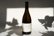 A bottle of 2017 Michael Mara Chardonnay from Broc Cellars sits on a flat surface, its label highlighting Sonoma Coast and Michael Mara Vineyard, while leaf shadows create an elegant pattern on the wall.