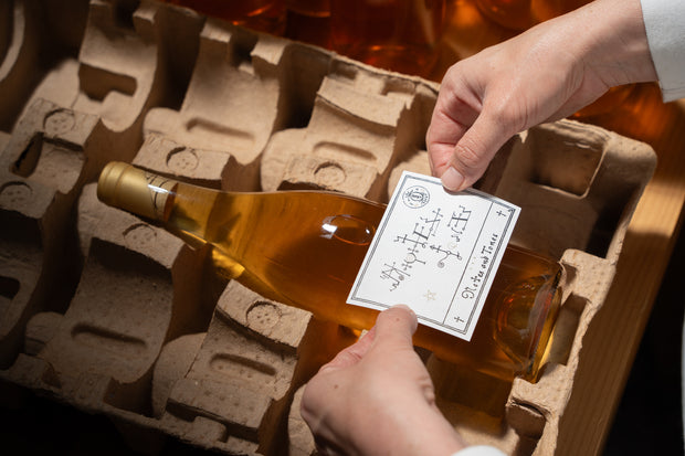 A person holding a Broc Cellars Notes & Tones V.2 bottle meticulously labels it. This collaboratively produced, organically farmed wine rests in cardboard crates along with more bottles of the same collection.