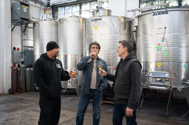 Three men are in Broc Cellars tasting the Notes & Tones V.2- The Album and Wine Set amidst large metal fermentation tanks. One holds a glass, another drinks deeply, while the third excitedly discusses flavors. The atmosphere is distinctly industrial.