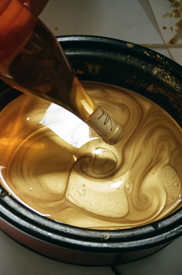A hand pours golden liquid from Broc Cellars Notes &amp; Tones V.2 - The Album and Wine Set into a black metallic pan, creating a swirling mix that gleams with organic artistry.
