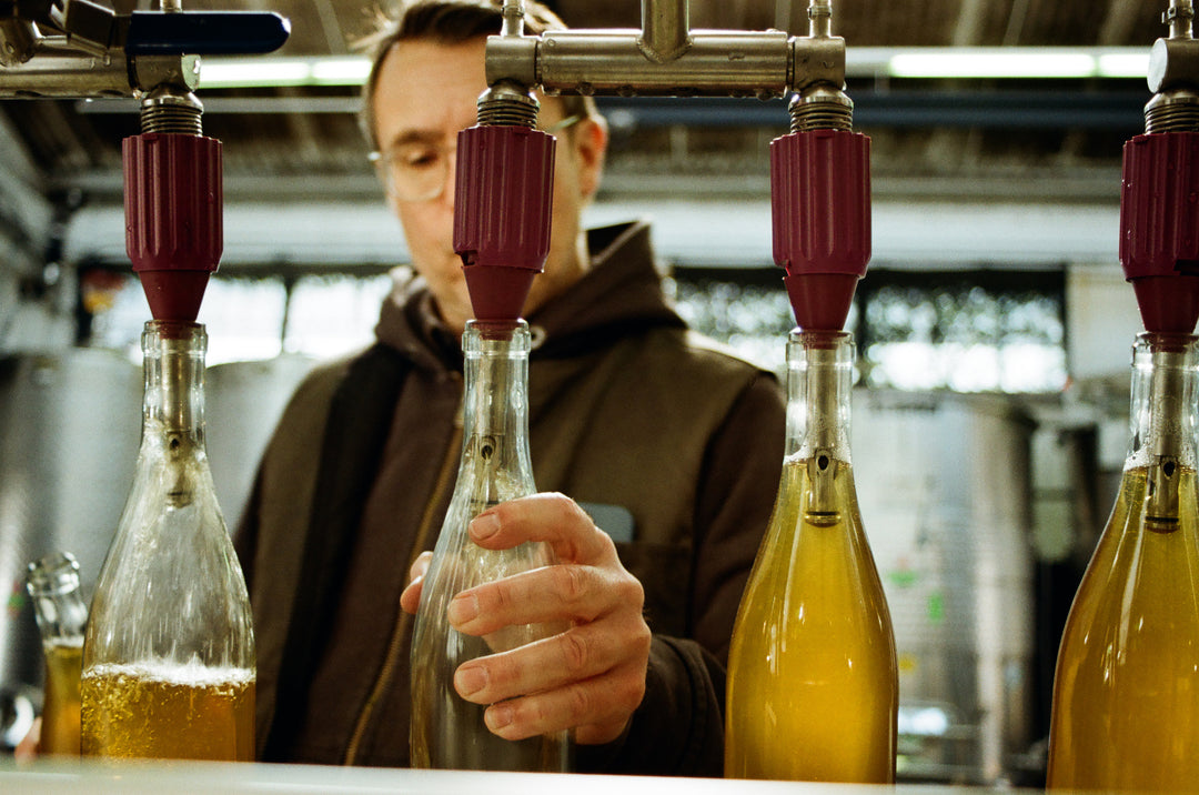 At Broc Cellars, the bottling machine fills four glass bottles with a yellow liquid, demonstrating their dedication to organic viticulture in an industrial facility. This setting underscores their commitment to collaboration on collections like Notes &amp; Tones V.2- The Album and Wine Set.