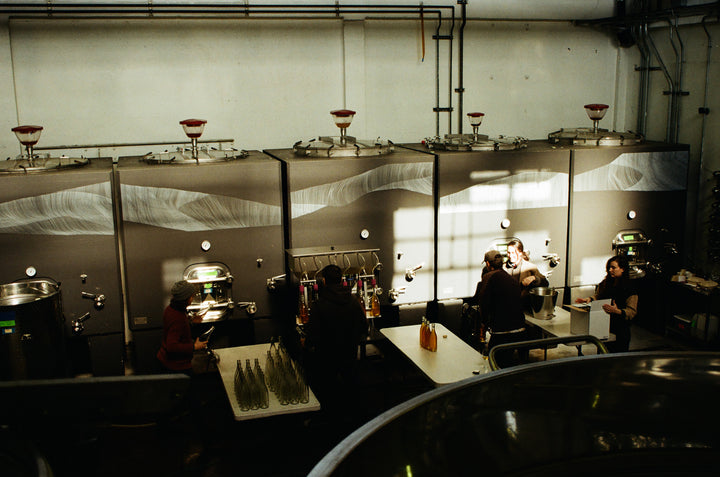 Inside an industrial facility, Broc Cellars crafts its collaborative wine, Notes &amp; Tones V.2- The Album and Wine Set. Workers operate near large metal tanks while tables display bottles and organically sourced supplies in a high-ceiling room lit by overhead lights and a window.