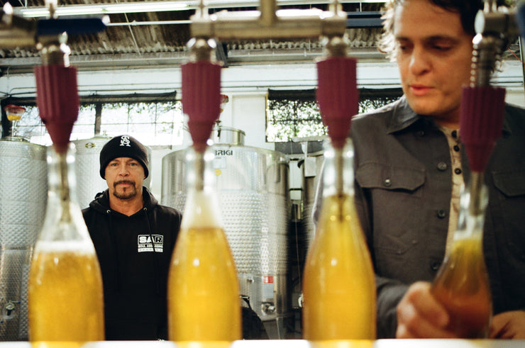 In a brewery, the man on the left in a black beanie and shirt stands by large Broc Cellars brewing tanks. The other man focuses on bottles being filled with organic wine from the Notes & Tones V.2 - The Album and Wine Set amid an industrial stainless steel backdrop.