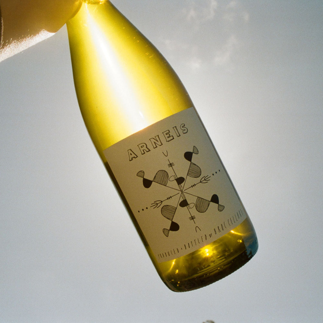 A hand holds a bottle of Arneis wine up to the sky, with sunlight shining through the yellowish glass. The label has a geometric black and white design and the word “ARNEIS” at the top.