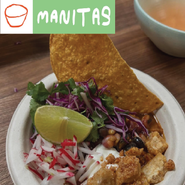 A bowl of food topped with tortilla chips, chopped cilantro, shredded purple cabbage, radish, lime wedge, beans, and crispy pieces, placed on a wooden table. The top left shows a green square with MANITAS text and a bowl icon.