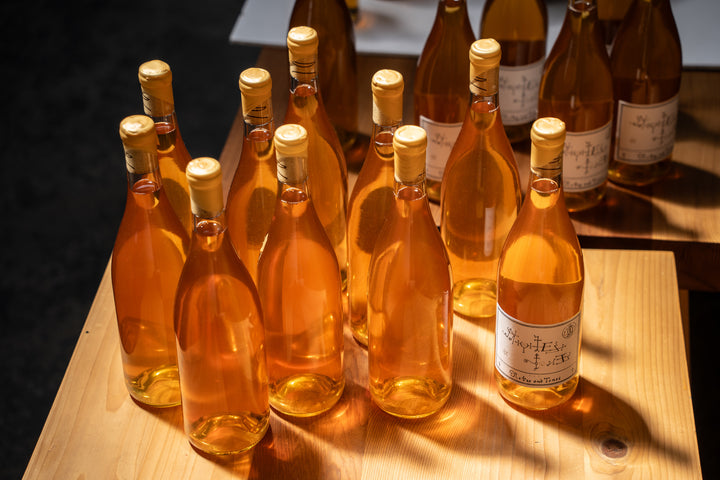 Several amber glass bottles with pale yellow labels and cork tops, from Broc Cellars Notes &amp; Tones V.2 - The Album and Wine Set, are arranged on a wooden table. Sunlight casts shadows, making some labels partially visible.