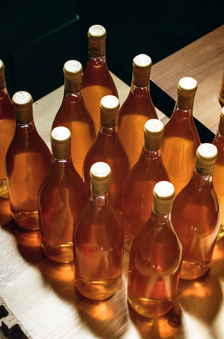 A group of glass bottles filled with amber liquid, sealed with beige caps, are neatly arranged on a wooden surface. They contain the unique Notes & Tones V.2 wine blend from Broc Cellars and beautifully catch the light in their surroundings.