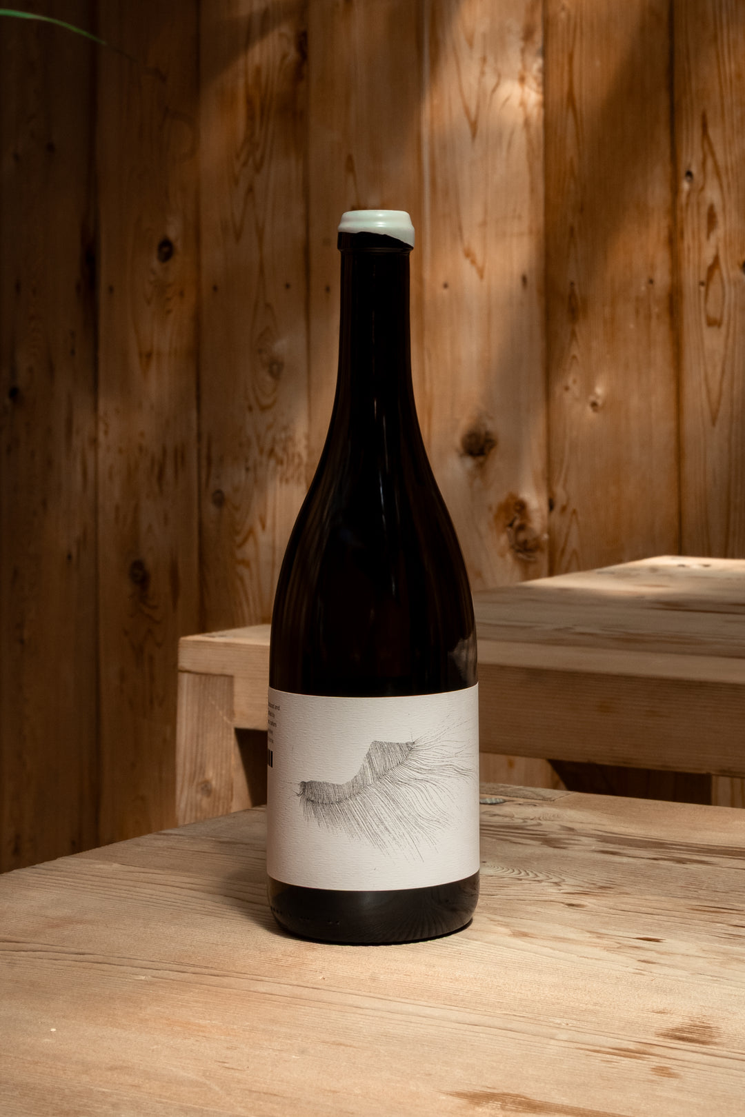 A dark glass bottle of 2016 MICHAEL MARA CHARDONNAY by Broc Cellars, with a white label and minimalist black line drawing, rests on a wooden table in a softly lit room with wooden walls.