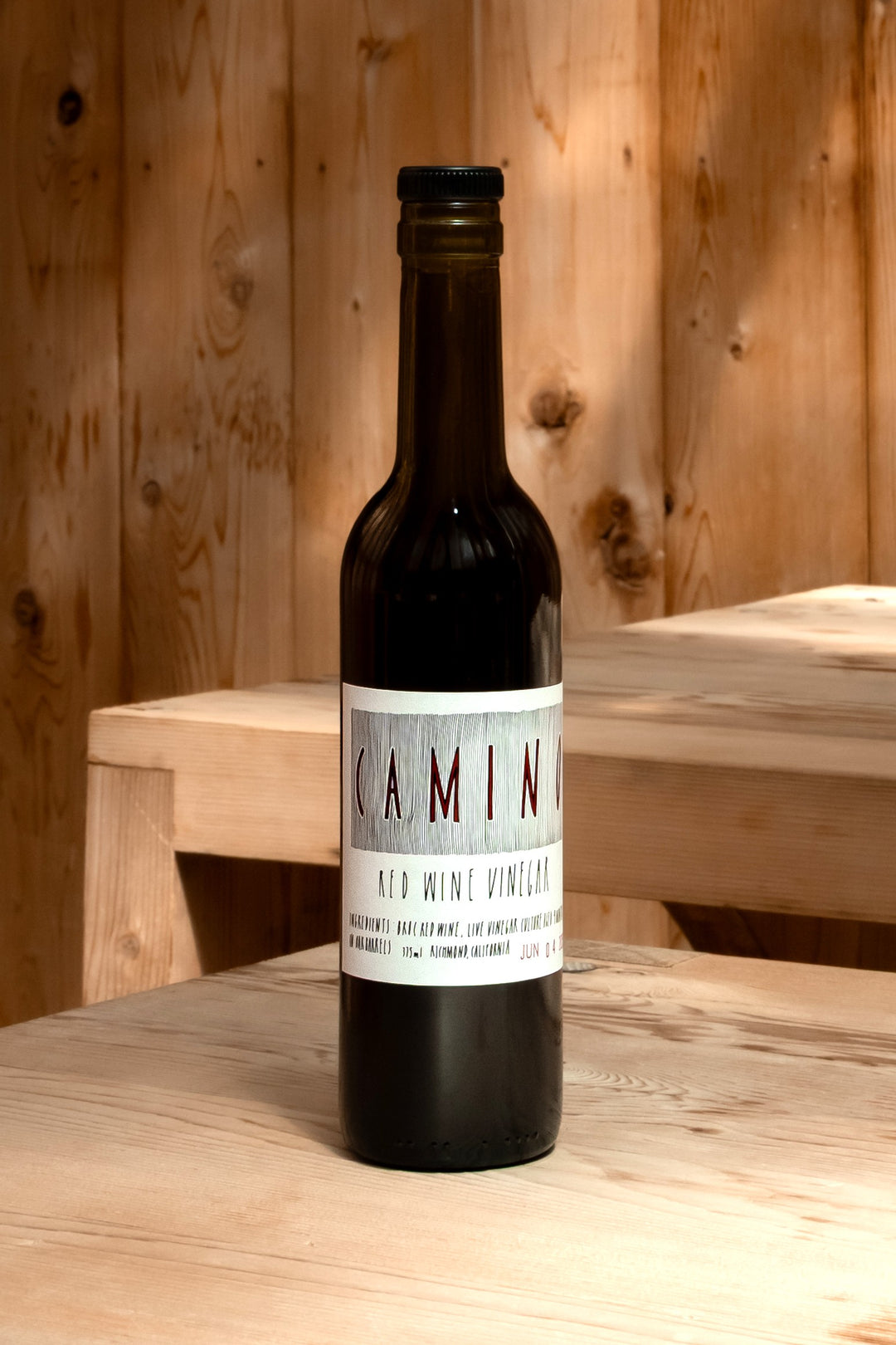 A dark glass bottle labeled Camino Red Wine Vinegar from Broc Cellars stands on a light wooden surface with a wooden wall and bench behind. Warm, natural light highlights this unique organic vinegar.